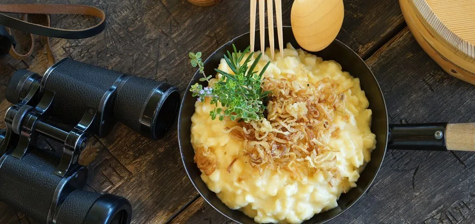 Käsknöpfle in einer gusseisernen Pfanne mit Röstzwiebeln, daneben ein Fernglas auf dem Holztisch