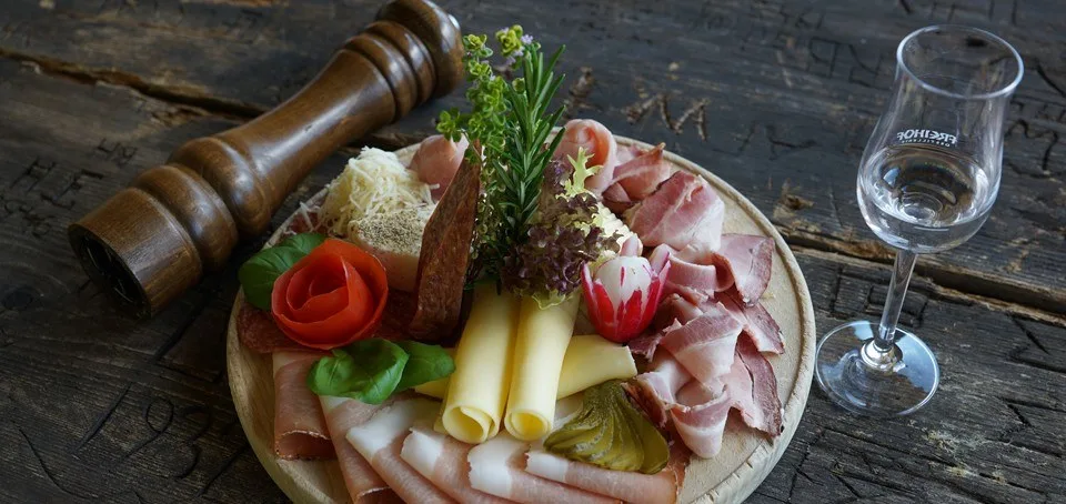 Brettljause mit Schinken, Bergkäse und Speck auf einem Holzbrett, daneben ein Stamperl