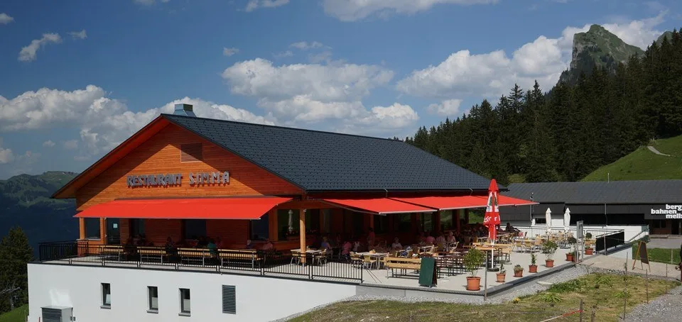 Das Bergrestaurant Simma von außen im Sommer, rote Sonnenschirme auf der Terrasse, dahinter die Bergkette