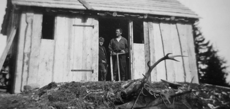 Historische Schwarzweißaufnahme: zwei Personen vor dem Holztor der alten Alphütte