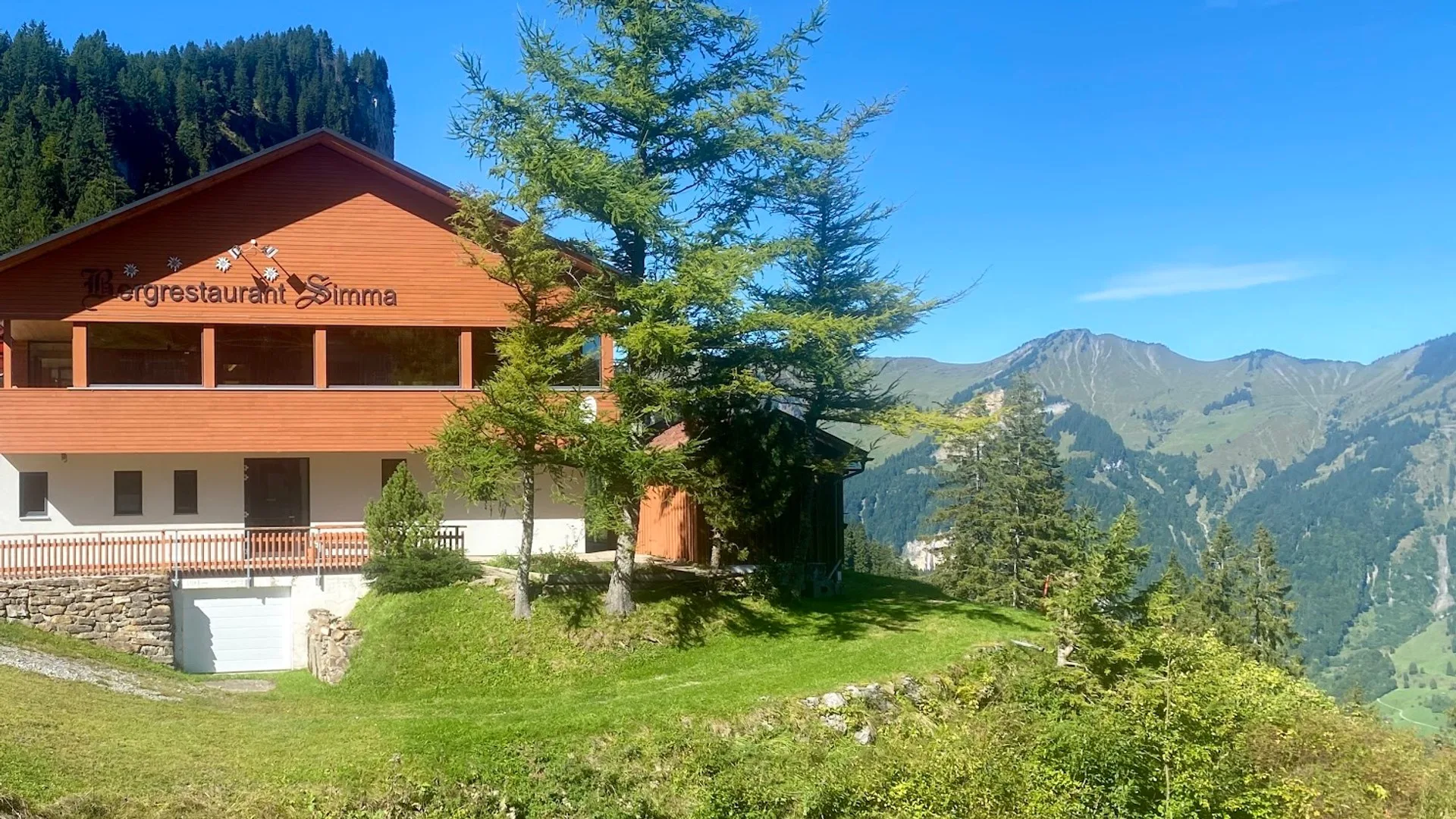 Das Bergrestaurant Simma auf der Rossstelle im Sommer, der Schriftzug am Giebel, davor Lärchen und dahinter die Bergkette